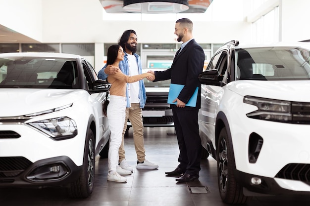 Um vendedor de carros profissional vestido de fato está apertando as mãos de um casal de clientes indianos felizes