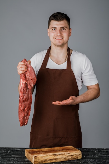 Um vendedor ou cozinheiro do sexo masculino segura carne crua nas mãos e a mostra sobre um fundo cinza.