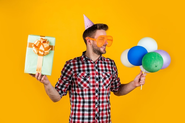 Um verdadeiro viciado em compras segura balões festivos, feliz feriado, tempo de celebração para se divertir e apresenta feliz aniversário ou aniversário, sentir alegria e alegria, puro festeiro, homem engraçado no boné de aniversário