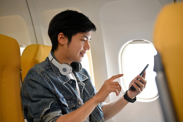 Um viajante asiático de boa aparência no assento da janela na classe econômica usando seu telefone celular