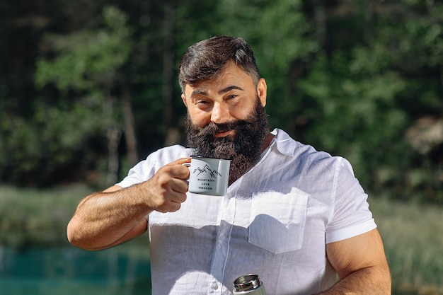 Um viajante brutal adulto com barba fica no contexto de uma floresta com árvores verdes e segura uma xícara com uma impressão e uma garrafa térmica nas mãos e sorri. beber uma bebida quente.