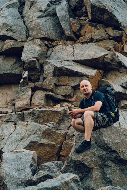 Um viajante com uma mochila atrás dos ombros senta-se na rocha ao pé de um penhasco alto.