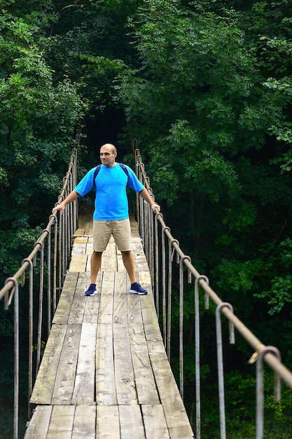 Um viajante masculino com uma mochila caminha ao longo de uma ponte suspensa sobre um rio de montanha