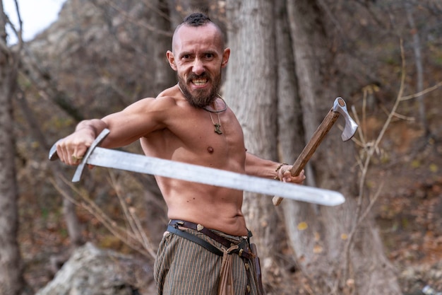 Foto um viking barbudo com uma expressão de raiva em uma posição de combate segurando uma espada e um machado de batalha