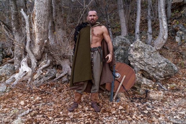 Um viking barbudo com uma pele de animal jogada sobre os ombros está de pé ao pé de uma montanha entre as árvores