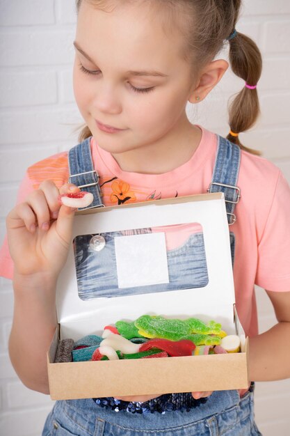 Uma adolescente bonita com duas tranças está segurando uma caixa inteira de marmelada multicolorida de formas engraçadas