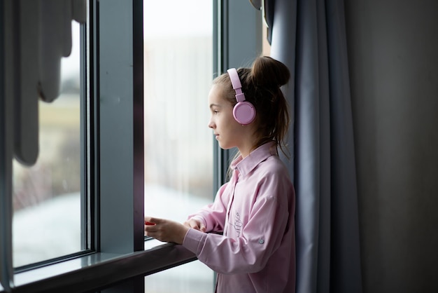 Uma adolescente bonita em uma camisa rosa e fones de ouvido olha pela janela e sonha.