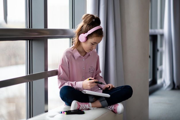 Uma adolescente bonita em uma camisa rosa e fones de ouvido senta-se perto de uma janela e desenha com marcadores. Criação.