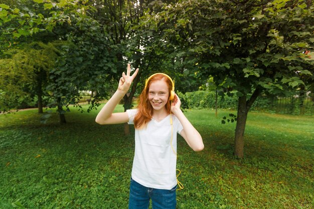Uma adolescente com cabelo ruivo ouve música com fones de ouvido em um parque no verão. Uma jovem aprecia e relaxa ao ar livre em um dia ensolarado.
