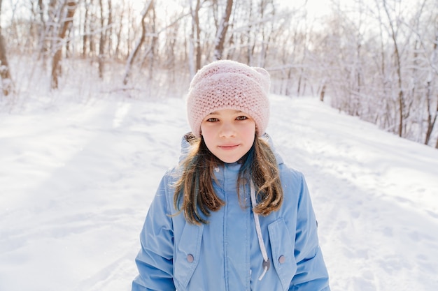 Uma adolescente com um casaco azul e um chapéu rosa está em uma floresta coberta de neve.