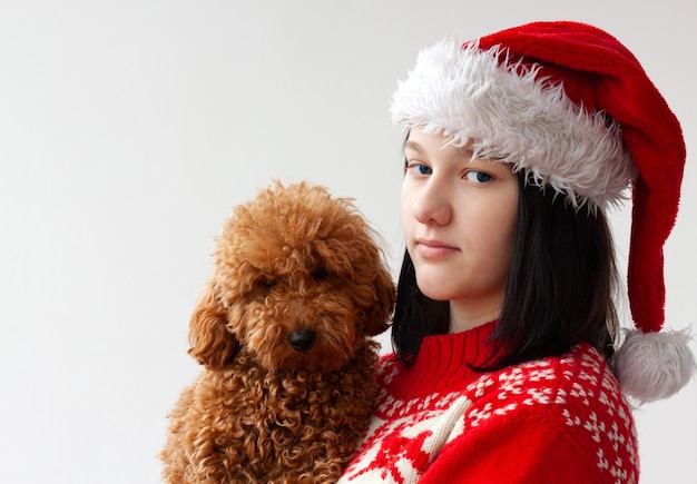 Uma adolescente com um chapéu de Papai Noel e um suéter vermelho de Natal segura um poodle miniatura nos braços. O conceito de Natal e Ano Novo