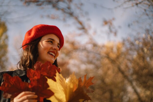 Uma adolescente com uma boina vermelha e um buquê de folhas de outono nas mãos caminha pela floresta