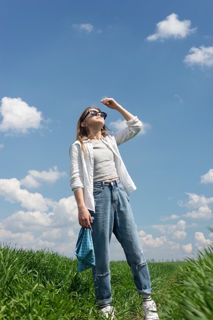 Uma adolescente de óculos escuros, em um campo de grama verde, se escondendo do sol, olha para cima, no contexto de um céu nublado.