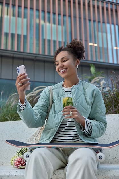 Uma adolescente feliz e despreocupada faz videochamada via smartphone, segura uma garrafa de bebida refrescante, senta-se com um skate sorrindo alegremente para a câmera, tira selfie em poses de retrato em ambiente urbano