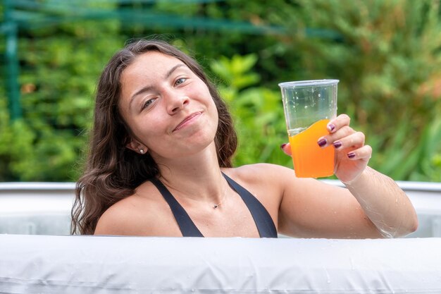 Foto uma adolescente linda com cabelos longos e escuros e um copo com bebida na piscina inflável do jardim.