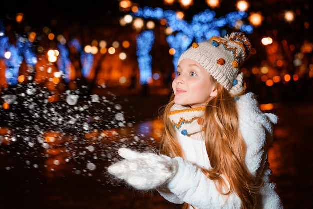 Uma adolescente loira com cabelo comprido em um chapéu e roupas de inverno em uma noite festiva de inverno na rua ...