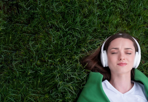 Uma adolescente ouve música em fones de ouvido, deitada na grama verde do parque, sorri e mostra