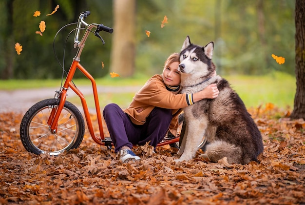 Uma adolescente se senta em uma scooter entre a folhagem do outono em um parque e abraça seu cachorro Husky Siberiano