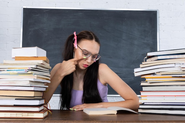 Foto uma adolescente senta-se na sala de aula entre as pilhas de livros e começa a ler a primeira leitura o aluno começa a aprender seu cabelo é preso com uma fita rosa em duas caudas de cavalo