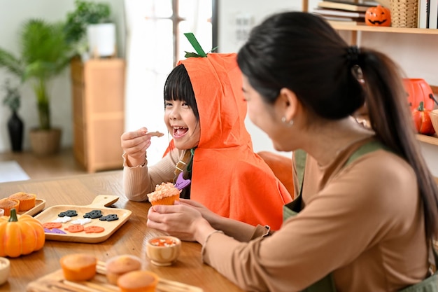 Uma adorável garota asiática em traje de Halloween gosta de comer cupcake com sua mãe