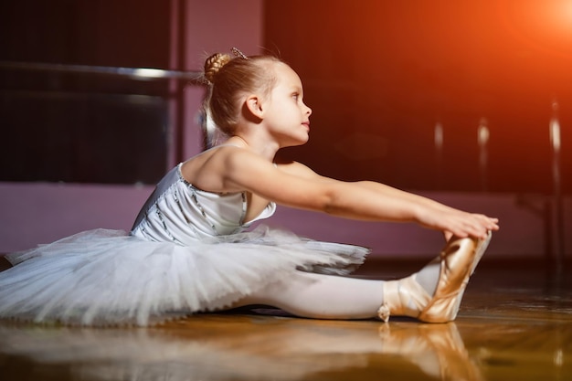 Uma adorável jovem bailarina de vestido branco sentado no chão de madeira e fundo de estúdio de dança Linda menina sonhando em se tornar dançarina de balé profissional escola de dança clássica