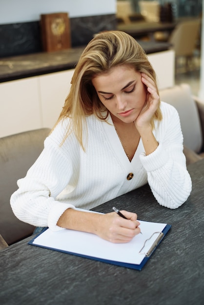 Uma adorável jovem está sentada à mesa em sua cozinha e escrevendo em um caderno.