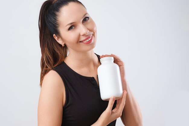 Foto uma alegre mulher caucasiana vestindo um top sem mangas preto segurando uma garrafa de suplemento branco contra um fundo claro que exala uma vibração amigável mockup para design