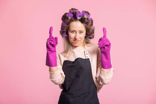 Uma alegre mulher sorridente usando luvas de segurança de borracha aponta para cima para um espaço vazio com espaço para publicidade Dona de casa no fundo rosa do estúdio