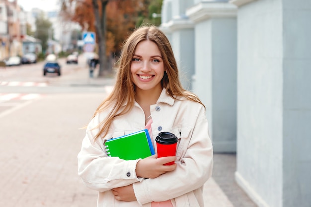 Uma aluna caminha pela cidade com uma xícara de café e um caderno. Rapariga estudante com cabelos brancos