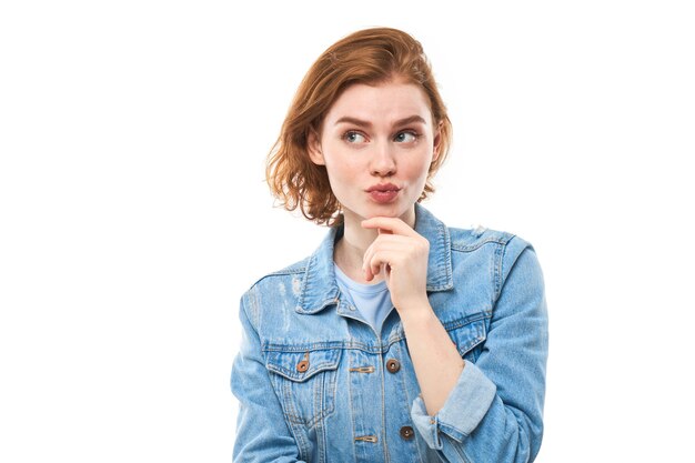 Uma aluna de cabelo vermelho feliz pensa e olha para a câmera enquanto segura o dedo perto do queixo. Mulher pensativa pensativa em uma jaqueta jeans azul sobre fundo branco. Copie o espaço