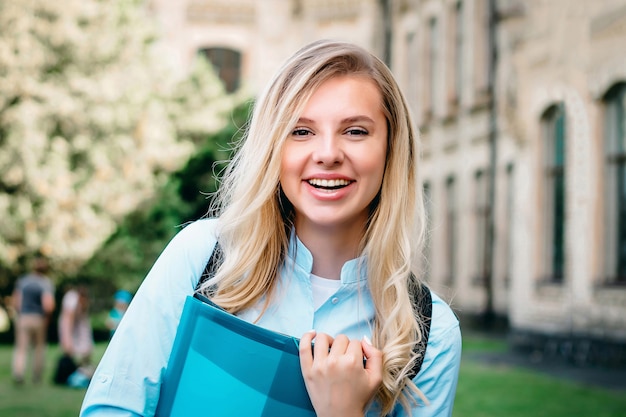 Uma aluna loira está sorrindo e segurando livros e um caderno nas mãos dela sobre um fundo universitário