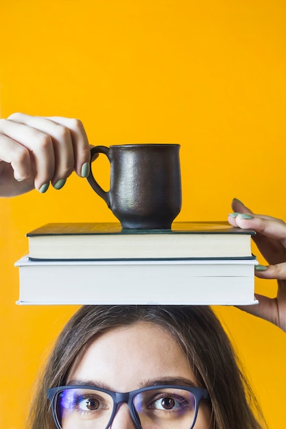 Foto uma aluna usando óculos está segurando uma pilha de livros e uma xícara de café na cabeça dela