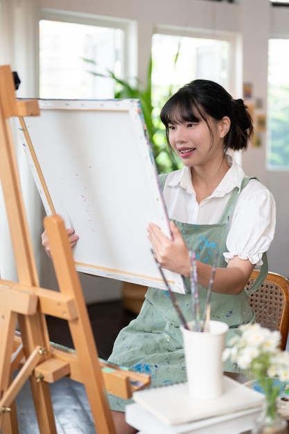 Uma artista asiática satisfeita segurando uma tela olhando para seu quadro pintado em seu estúdio