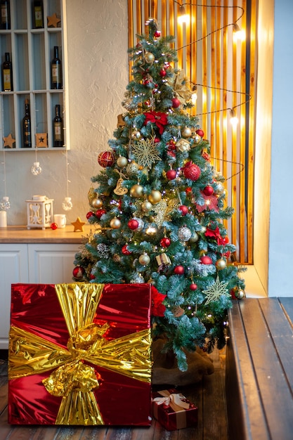 Uma árvore de natal decorada com um enorme presente embaixo dela. foto vertical. grande caixa brilhante de croas com fita sob a árvore de natal no interior da casa