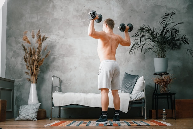 Uma atleta com cabelos loiros balança os bíceps em halteres no quarto, treinamento online. O jovem pratica esportes em casa.