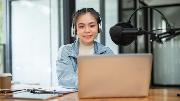 Uma atraente apresentadora de rádio asiática está olhando para a tela de seu laptop falando em um microfone