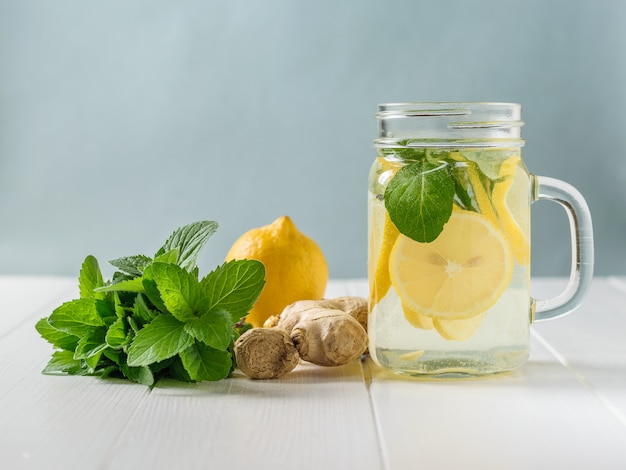 Uma bebida refrescante de limão, hortelã e fatias de raiz de gengibre em uma mesa de madeira branca.