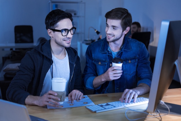 Uma bebida saborosa. Lindos programadores positivos rindo e desfrutando de seu café enquanto estão sentados na frente do computador