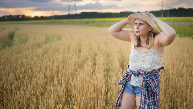 Uma bela agricultora de meia-idade com um chapéu de palha e uma camisa xadrez fica em um campo de trigo maduro dourado durante o dia à luz do sol