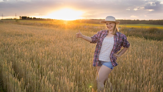 Uma bela agricultora de meia-idade com um chapéu de palha e uma camisa xadrez fica em um campo de trigo maduro dourado durante o dia à luz do sol