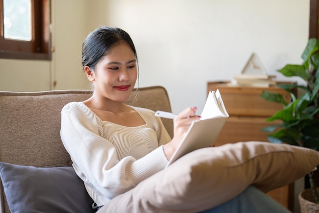 Uma bela e feliz mulher asiática está mantendo seu diário enquanto relaxa em um sofá em sua sala de estar