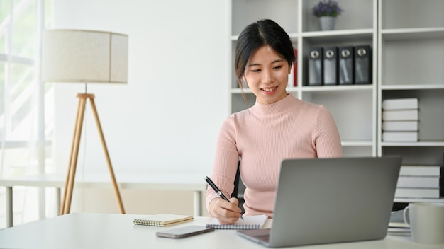 Uma bela empresária asiática se concentra em seu trabalho fazendo anotações e usando seu laptop