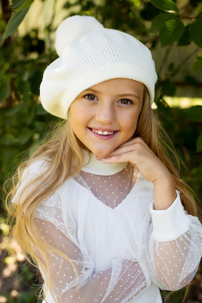 Foto uma bela garota loira feliz em uma blusa branca e um chapéu com um pompom está de pé na rua