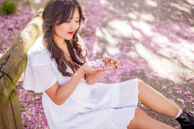 Uma bela jovem asiática em um vestido branco caminha em um parque florido. Sakura. Árvores florescendo. Primavera.