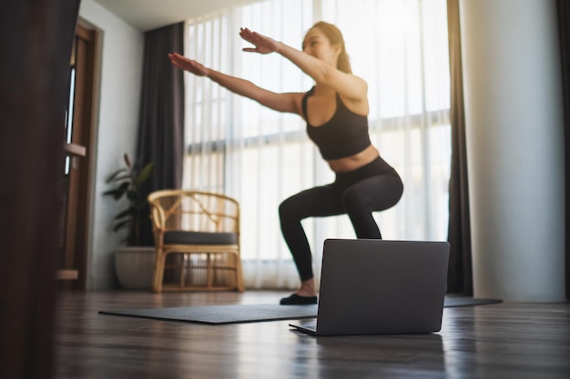 Uma bela jovem asiática fazendo agachamentos enquanto assiste a tutoriais de exercícios on-line no laptop em casa