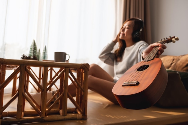 Uma bela jovem asiática segurando um ukulele e gosta de ouvir música com fone de ouvido em casa