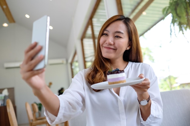 Uma bela jovem asiática usando telefone celular para tirar uma selfie com um bolo antes de comer no café