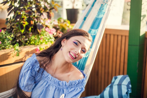 Uma bela jovem com maquiagem brilhante e longos cabelos escuros está sentado em uma rede no terraço de verão de um café da cidade, olhando para a câmera e sorrindo