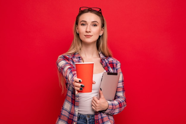 Uma bela jovem com roupas elegantes segura um tablet e um copo de papel para café em um fundo vermelho