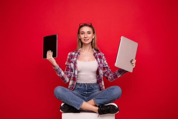 Uma bela jovem com roupas elegantes segurando um tablet e um laptop em um fundo vermelho